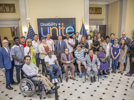 A large group of advocates and elected officials pose beneath a “Disability Unite” banner