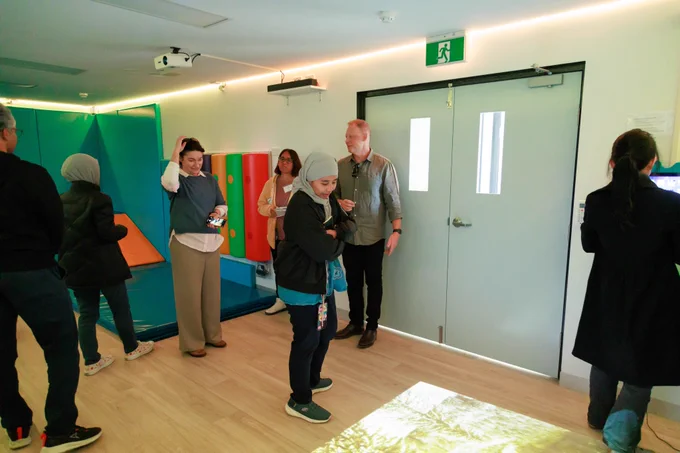 A group of adults explore a sensory room equipped with padded walls, colorful sensory equipment, and interactive floor projection.