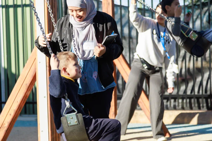 An adult wearing a hijab smiles warmly while assisting a young child sitting on a playground swing outdoors.