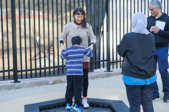 An adult happily supports a child standing on a trampoline, while others observe in an outdoor playground area.