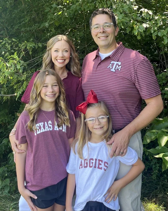 Family of four wearing Texas A&M University themed apparel, smiling outdoors with green foliage in the background.