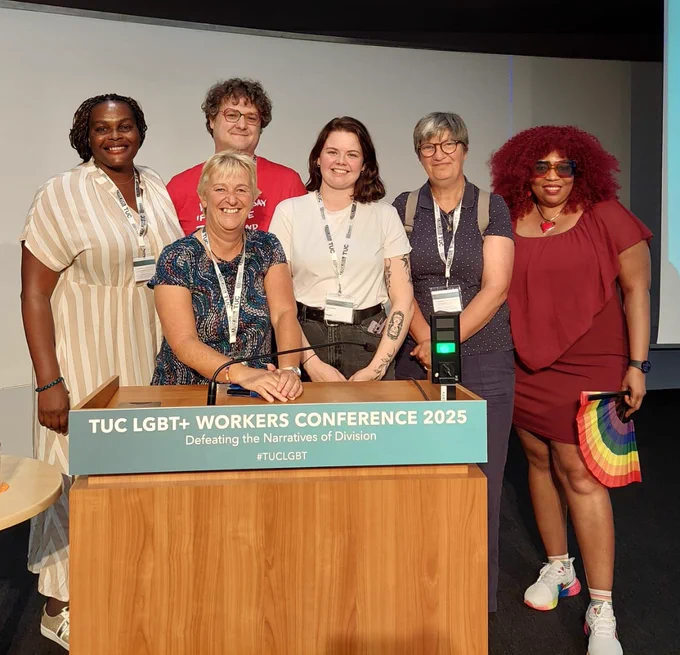 6 people standing together behind the TUC LGBT Conference lectern, all smiling at the camera and looking happy and positive.