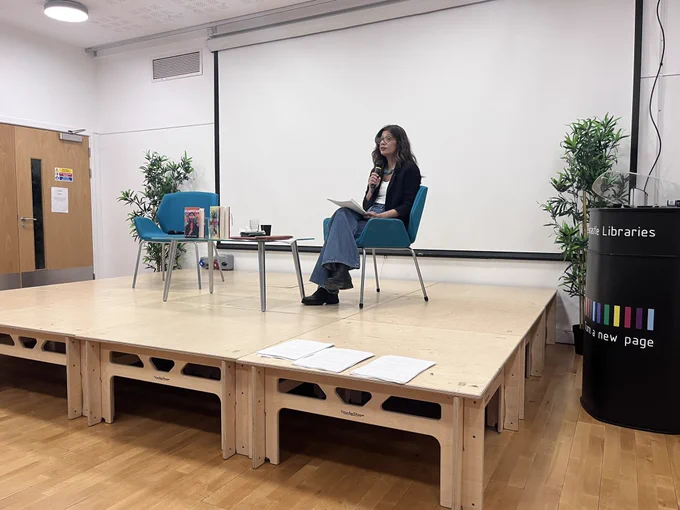 Sabyn Javeri, sitting on the stage in Newcastle City Library. She has dark hair and is wearing blue jeans and a black blazer. She is also wearing dark-rimmed glasses.