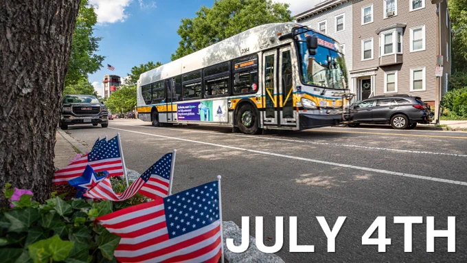 MBTA bus driving past a street decorated with small American flags and flowers on a sunny July 4th day.