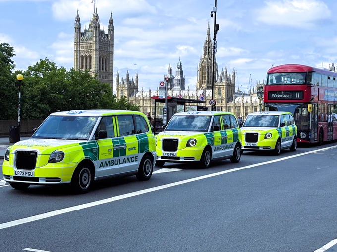 Three white taxi-ambulances drive south across Westminster Bridge. They are yellow and green marked as Ambulance. There is a red London bus behind them, and the Houses of Parliament in the background.