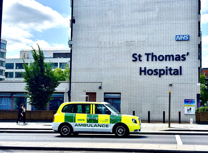 A white ambulance-taxi with yellow and green ambulance markings and the Trust logo drives past the side of St Thomas' Hospital, onto Westminster Bridge Road