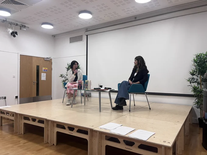 Sabyn Javeri, sitting on the stage in Newcastle City Library. She has dark hair and is wearing blue jeans and a black blazer. Yeisil is sitting with her, who is wearing a white shirt, a dark top and a skirt with a cherry pattern on it. She has dark hair and is wearing clear-rimmed glasses.
