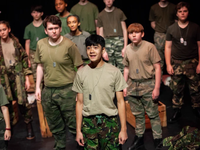 Teenagers in military costumes perform on a theatre stage.
