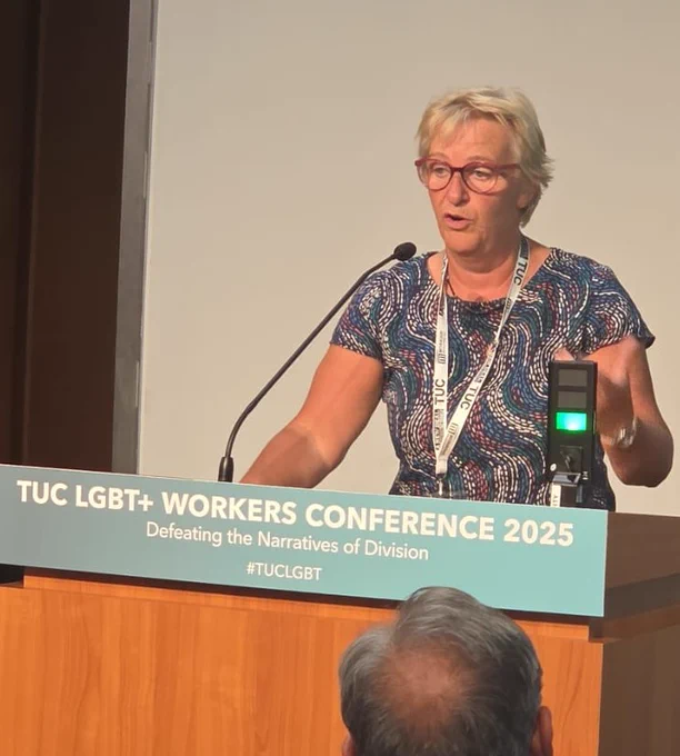Jackie has short blonde hair and wears glasses and a patterned top. She is standing and speaking at the TUC LGBT  Workers Conference lectern.