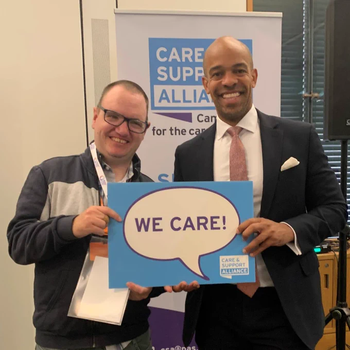 A photo of Sean Nightingale and MP Ben Obese-Jecty. They are both smiling. They are standing in front of a Care and Support Alliance banner and holding a sign which reads: We Care!