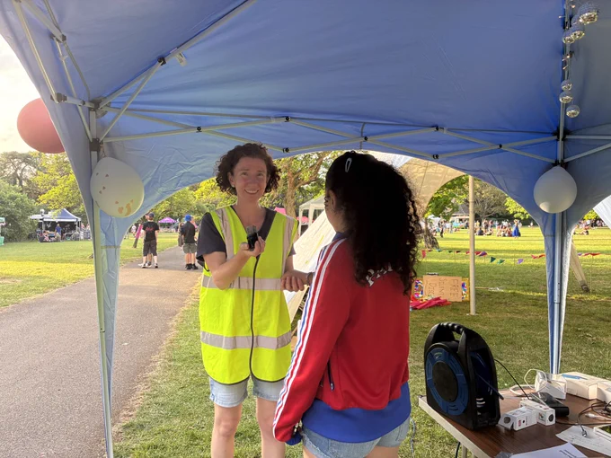 Anneliese wearing a high viz vest, stood under a gazebo with a park in the background, in the process of stamping the hand of a young person with an entrance stamp.