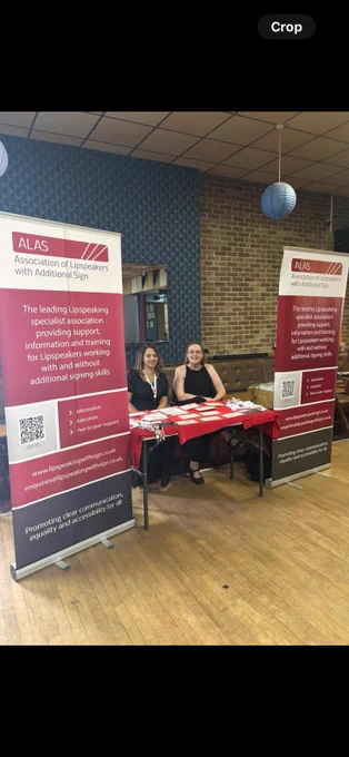 A picture of two women sat at a table. There are two banners either side of the table that read ‘ALAS Association of Lipspeakers with Additional Sign’