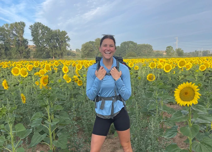 A person with a backpack stands smiling in a vast field of sunflowers on a sunny day.