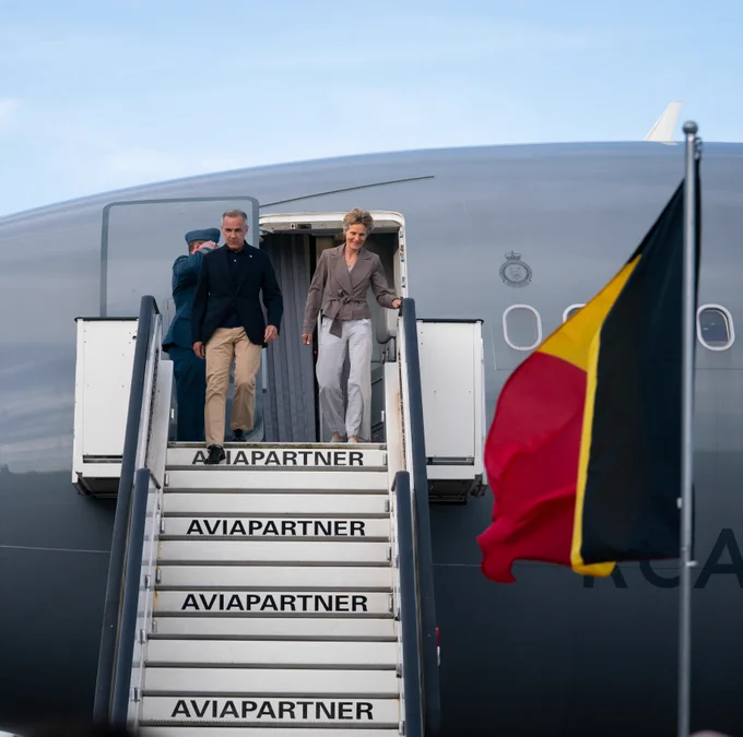 Prime Minister Mark Carney and Diana Fox Carney walk down the steps of a Royal Canadian Air Force aircraft. A Belgian flag is in the foreground.