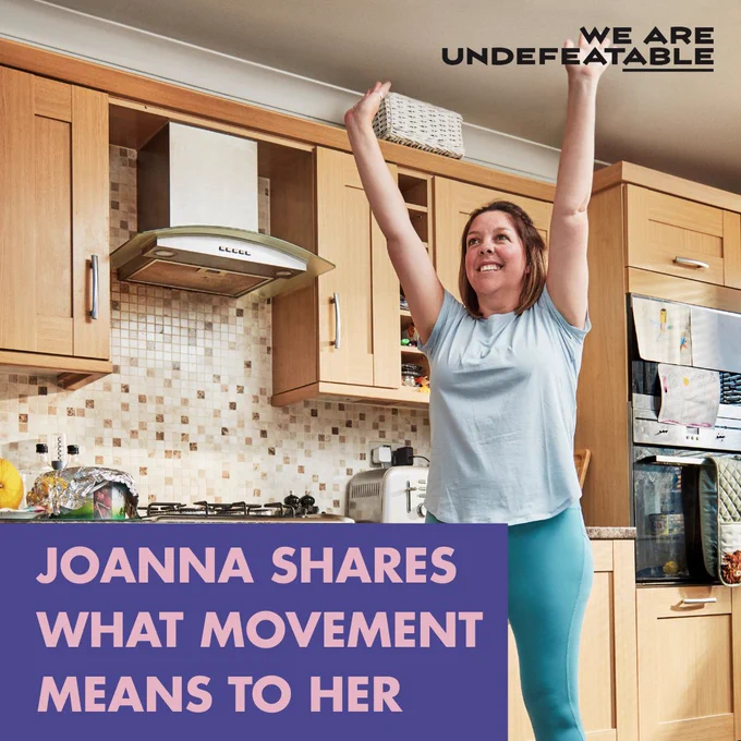 Woman excitedly stretches her arms up in a kitchen,  text reads 'Joanna shares what movement means to her' with a campaign logo 'We Are Undefeatable' displayed.