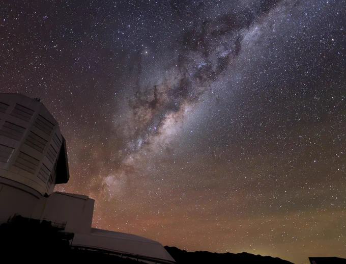 The glowing band of the Milky Way emanates from Rubin Observatory's boot-shaped silhouette at lower left. The brighter, dense stars of the Milky Way are crisscrossed by clouds and lanes of dark dust. The lower left horizon has an orangey hue, while the pinpricks of stars speckle the rest of the sky.