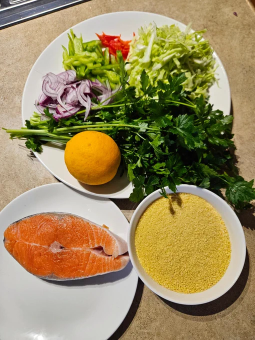 Ingredients clockwise from top of a round white plate with thinly sliced red chilli, cabbage, green capsicum, red onion, a handful of flatleaf parsley, a lemon. A small bowl of dry couscous, a small plate with a salmon steak.