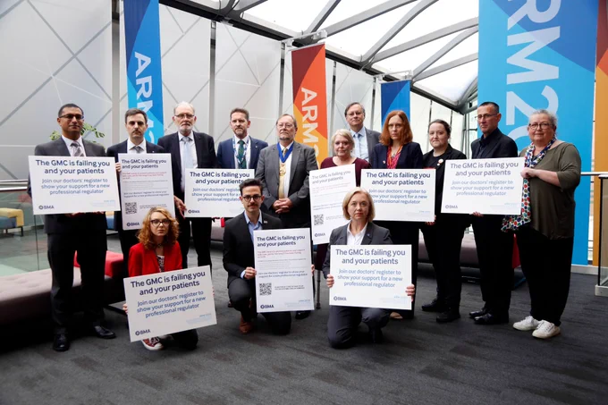 A group of BMA doctors are photographed holding signs that say, 'The GMC is failing you and your patients. Join our doctors' register to show your support for a new professional regulator.' In the background a conference venue including ARM2025 banners can be seen.