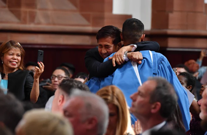 A man warmly hugs his son, who is at a graduation ceremony wearing a graduate's academic gown. The graduate's mum is using her smartphone to take a photograph of the happy pair.