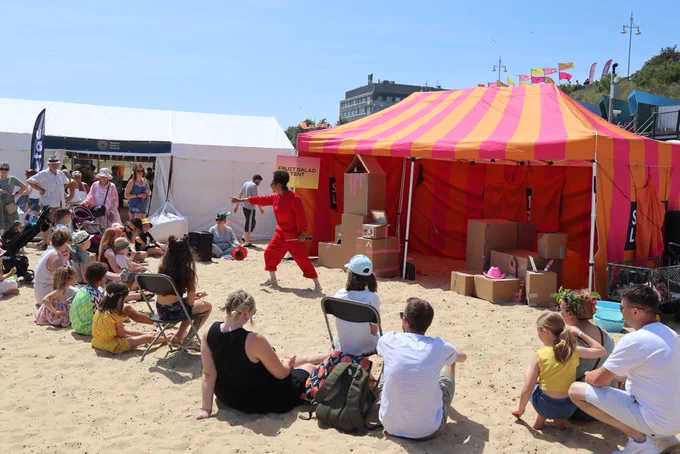 Performer points to the audience sitting on the beach in front of the performance tent / stage.