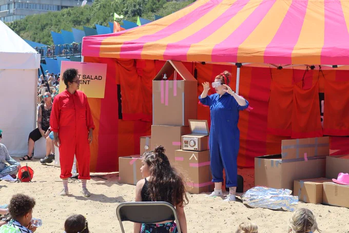 Two performers on the beach performing in front of cardboard props and wearing bright jump suits
