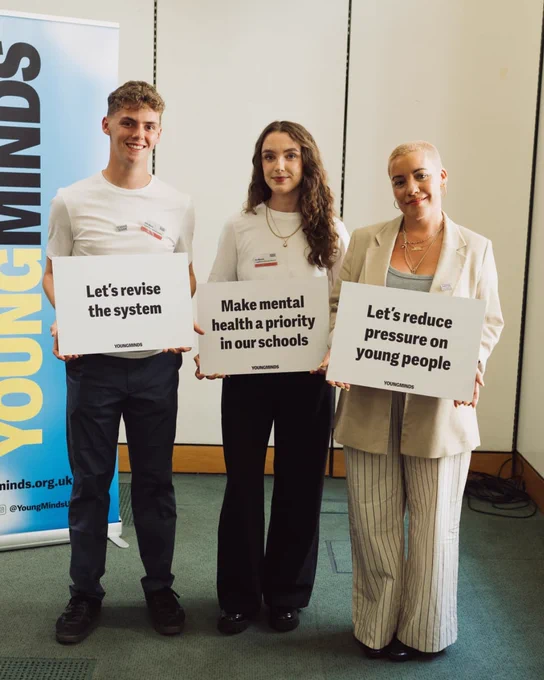 Our Ambassadors Alfie Watts (left), Ro Mitchell (middle), and Gemma Ogston holding up signs infront of a Young Minds billboard. Alfie's sign says 'Let's revise the system', Ro's sign says 'Make mental health a priority in our schools' and Gemma's sign says 'Let's reduce pressure on young people'.