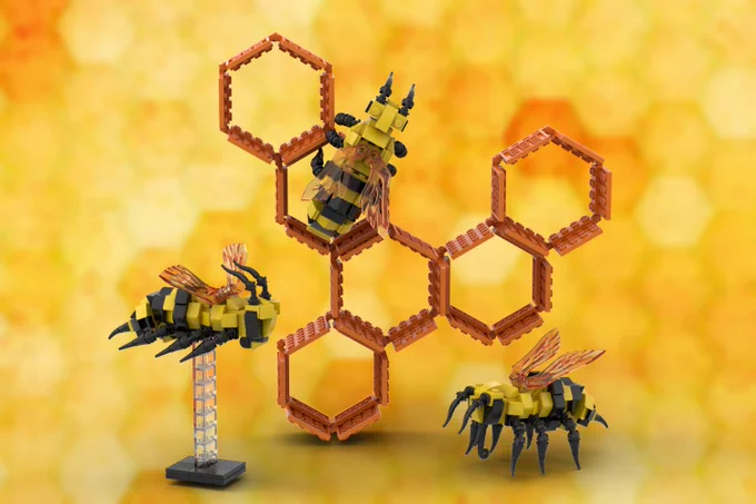Prototype LEGO bees and honeycomb showing 3 bees, each having a yellow body with black stripe, black, articulated legs, black antennae and mouth parts and amber wings.  One bee (left, foreground) is on a clear plastic LEGO plinth; a second bee (left of centre) is facing away from us on the an upper hexagon of a 6-hexagon, amber LEGO honeycomb (shaped roughly in a Y-formation, tilted slightly to the right); and the 3rd been on the 'ground', facing left, to the right side of the 'honeycomb'.  ALl against a yellow/amber background having feint hexagon watermarks all over. (phew!)  