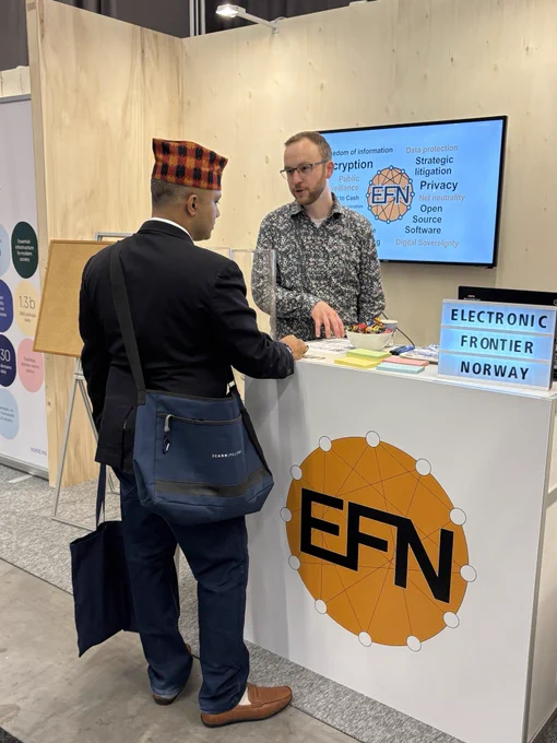 A booth at a conference with a table, screen and some posters. In front of the table is the logo of EFN, on the screen are topics EFN works with, like encryption and privacy