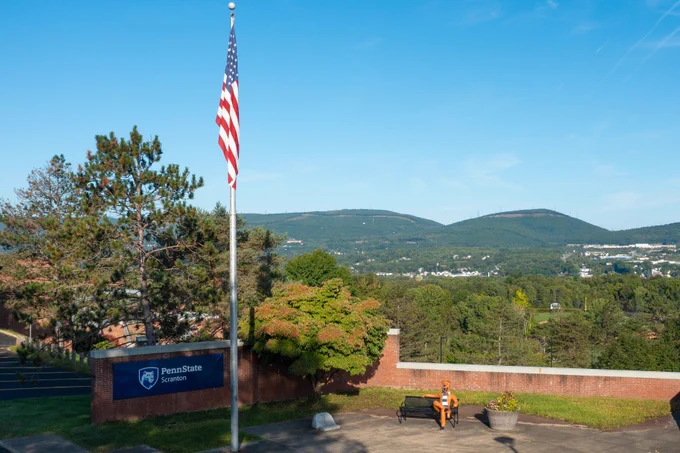American Flag in foreground at Penn State Scranton's campus, with Nittany Lion bench and 'Penn State Scranton' signage in background.