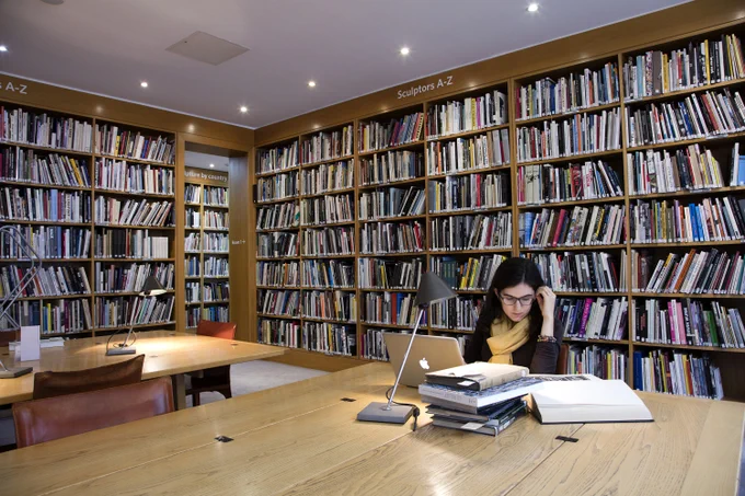 A woman sits studying at a desk in the library surrounded by shelves of books, working at a laptop with books piled on the table in front of her
