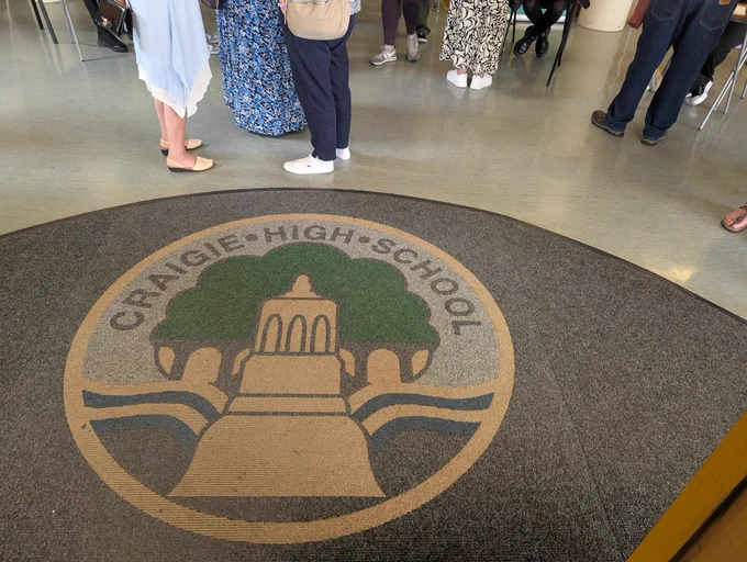 The Craigie High School logo on a semi circular carpet. It is a picture of a tower on a hill in front of a tree. There are wavy lines either side of the tower. At the top of the photo the legs of people can be seen standing in small groups. The photo does not show anyone above waist height.