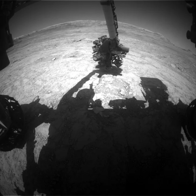 A black-and-white image taken by the Curiosity rover shows its own shadow cast on the rocky Martian surface. The shadow clearly outlines the rover’s body, mast, and robotic arm, which is extended over the ground. The barren terrain stretches out to the horizon, with scattered rocks and a slightly elevated ridge in the distance.