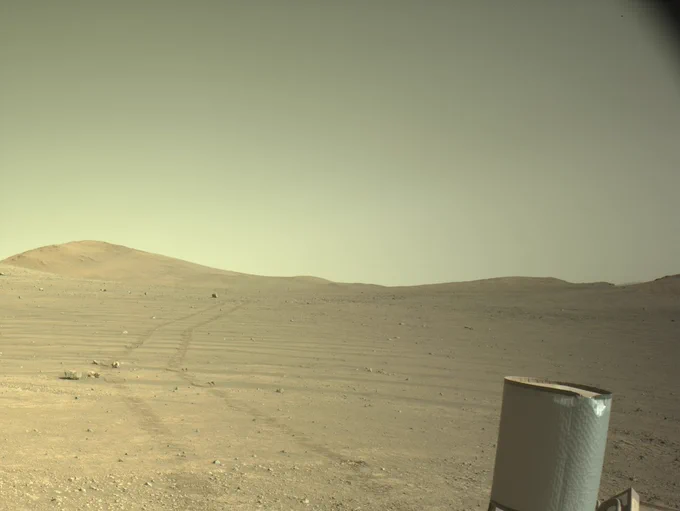 A view of rover tracks in the sandy Martian surface is shown in a photo taken by the Perseverance rover. The tracks go off into the hazy horizon and the image is tinted a beige color. A small piece of the rover is shown off to the lower right.