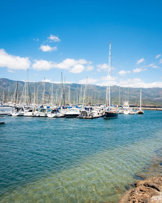 Santa Barbara Harbor  featuring multiple sailboats docked in calm waters. Clear blue sky overhead with some clouds, and mountains visible in the background.