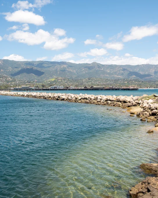 A serene waterfront scene featuring clear blue water and a the breakwater in Santa Barbara. Mountains visible in the background under a partly cloudy sky.