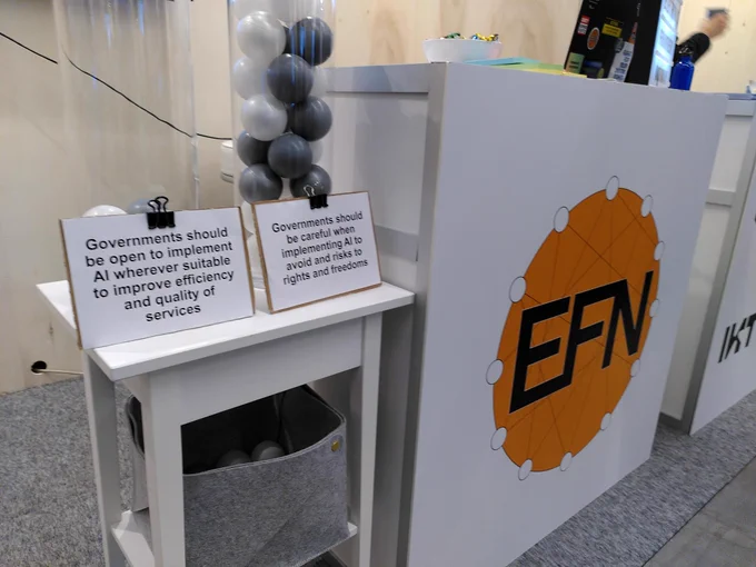 A section fo a stand. In the back you see a table with the logo of EFN at the front. In the front you see a table and two vases with balls that indicate votes. One vase is vuller than the other. The fuller vase says "Governments should be careful when implementing AI to avoid risks to rights and freedoms" the more empty vase says "Governments should be open to implement AI wherever suitable to improve efficiency and quality of services"