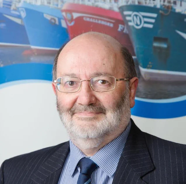 Ross Dougal, a man with a balding head, glasses and beard wearing a suit, stands in front of a poster with boats on it.