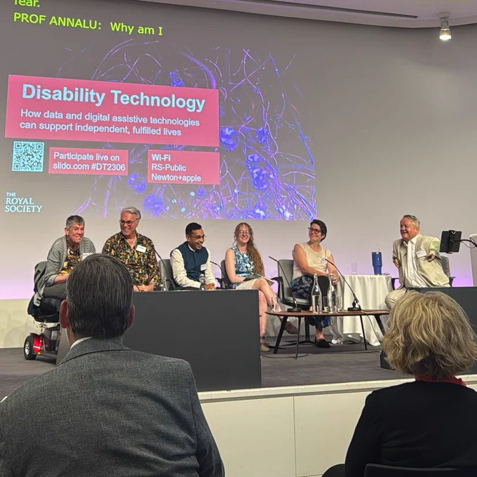 A wide shot shows a panel discussion on "Disability Technology" at the Royal Society. Six panelists are seated on a stage, facing an audience. The presenter, standing on the left, is speaking into a microphone. A large screen behind the panelists displays the title "Disability Technology" and the subtitle "How data and digital assistive technologies can support independent, fulfilled lives." The audience is visible from behind, with several people seated in the foreground.