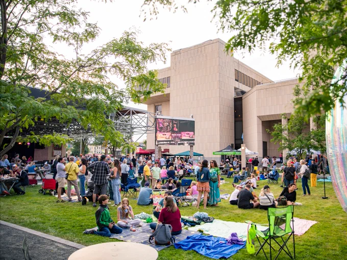 Outdoor event at the Marcus Performing Arts Center with people sitting on blankets, children playing, and food trucks in the background.