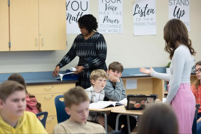 A teacher stands in the back of a classroom, slipping the pages of a book. Another teacher speaks to students as they listen.
