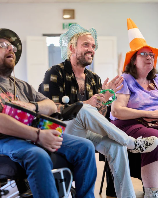 Three individuals participating in a festive event, with one person wearing a bright orange traffic cone as a hat and the others in playful attire, sitting together and enjoying a lively discussion.