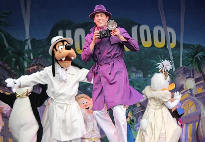 Performer in purple coat poses with camera alongside Goofy and Daisy Duck on a Hollywood-themed stage.