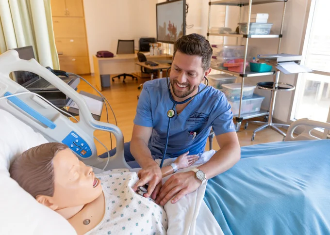 Nursing student smiles while practicing on a medical mannequin in a simulation room.