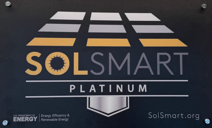 Plaque displaying the SolSmart Platinum designation. At the top is a stylized solar panel graphic in shades of gray and gold. Below, large text reads “SolSmart” with a sunburst design in the “O.” Underneath, a silver bar contains the word “Platinum.” In the lower left, it reads “U.S. Department of Energy, Energy Efficiency & Renewable Energy.” In the lower right: “SolSmart.org.”
