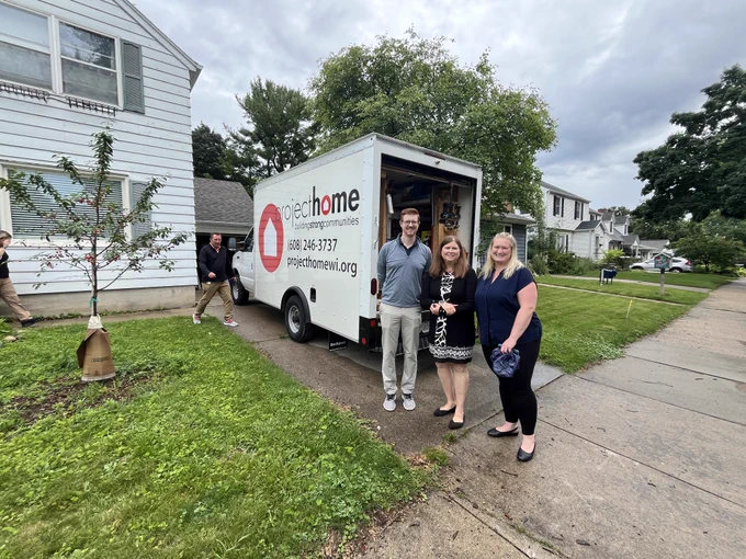 Secretary Blumenfeld, Assistant Deputy Secretary Maas, and DEHCR Deputy Administrator Mark Staff stand for a photo in front of a Project Home truck at a Project Home weatherization site house. The truck behind the group reads, "Project Home: Building Strong Communities" with a red house graphic and the organization's contact information.
