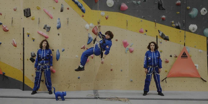 three women who look alike wearing blue jumpsuits at a climbing wall - two are standing on either side of the third who is hanging from a rope. A blue cat is standing on the ground to the left hand side.