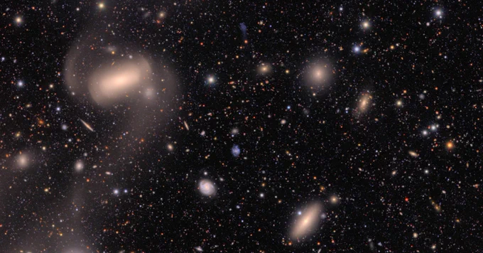 A field of galaxies of many shapes, sizes and colors, as well as the bright multi-colored points of stars. At top left is a large galaxy that is smooth like a stone, with wisps like cotton candy spiral from left and right. In the background, hundreds of galaxies of many shapes are just smudges of various colors against the black of space.