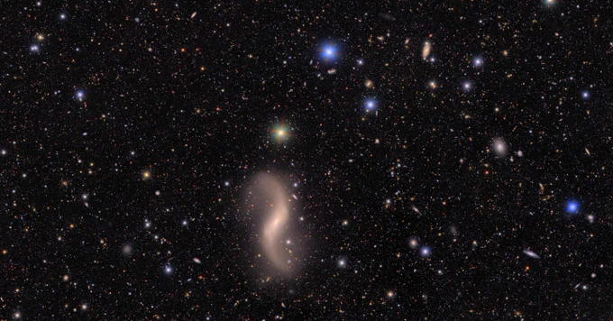 A field of galaxies of many shapes, sizes and colors, as well as the bright multi-colored points of stars. At bottom left is an elegant two-armed galaxy, with arms extended like a dancer viewed from above. The bright points of three stars form a triangle at top center. In the background, hundreds of galaxies of many shapes are just smudges of various colors against the black of space.
