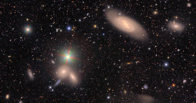 A field of galaxies of many shapes, sizes and colors, as well as the bright multi-colored points of stars. At top right is a large galaxy with two whirling spiral arms wrapping around like a hug. At bottom left, two smooth elliptical galaxies appear near each other, like champagne glasses in the midst of a cheers. A bright star above the pair has red, green, and blue diffraction spikes emanating out. In the background, hundreds of galaxies of many shapes are just smudges of various colors.