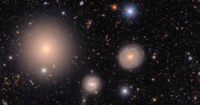 A field of galaxies of many shapes, sizes and colors, as well as the bright multi-colored points of stars. At left is a large elliptical galaxy, smooth like a polished stone. At right lies a large spiral galaxy that is nearly a ring, with bright golden core. To its lower left is a smaller spiral with a barred core. At top and bottom, two bright stars shine brightly with multi-colored diffraction spikes emanating out. In the background, hundreds of galaxies are just smudges of various colors.